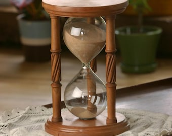 May include: A wooden hourglass with a sand timer inside. The hourglass is on a white lace doily on a dark surface.
