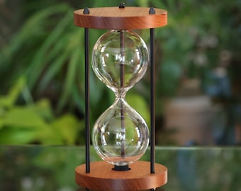 May include: A wooden hourglass with a black metal frame and two clear glass bulbs. The hourglass is sitting on a glass surface.