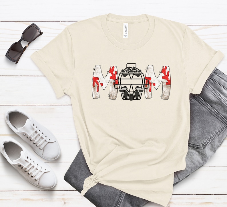 Baseball Shirt features distressed font spelling the word Mom and a Catcher's mask in place of the "o" in Mom.