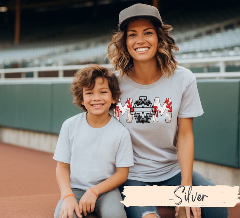 Baseball Shirt features distressed font spelling the word Mom and a Catcher's mask in place of the "o" in Mom.]