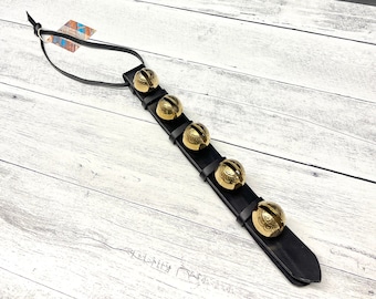 May include: A black leather strap with five gold jingle bells attached. The bells have a decorative design and are evenly spaced along the strap.