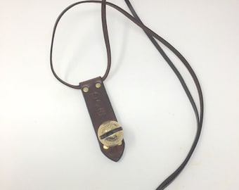 May include: A brown leather necklace with a pendant. The pendant has a circular brass design with a screw detail. The leather strap is knotted at the end. The pendant is attached to a rectangular leather piece with brass rivets.