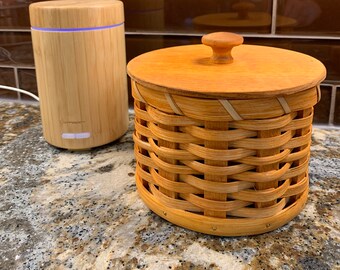 May include: A wooden essential oil diffuser with a bamboo exterior and a white light. The diffuser is sitting on a granite countertop next to a woven basket with a wooden lid.