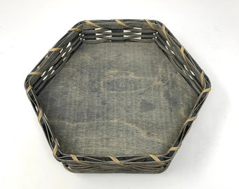 May include: Hexagonal wooden basket with a gray interior and woven sides. The basket has a light brown woven trim around the edges. The basket is empty and sits on a white surface.