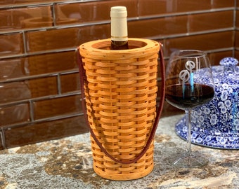 May include: A woven wicker wine bottle cooler with a brown leather handle. The cooler is holding a bottle of wine with a cork.