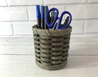 May include: A gray woven wicker pencil holder with blue pens and blue scissors inside.
