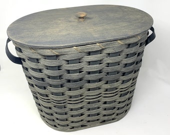 May include: A gray and black woven basket with a matching oval lid and a small wooden knob. The basket has black handles and is designed for storage. The basket is made of woven material.