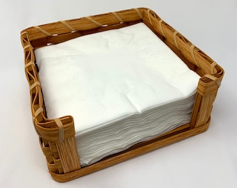 May include: A woven wicker napkin holder with a stack of white paper napkins inside.
