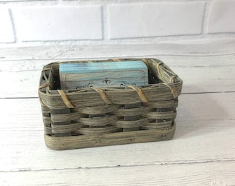 May include: A small woven basket with a light brown and gray pattern. The basket is filled with blue and brown cards. The basket is sitting on a white wooden surface.