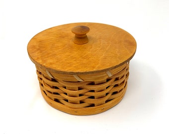 May include: A round, woven basket with a wooden lid and knob. The basket is made of natural wood and has a light brown color.