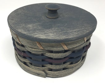 May include: A round woven basket with a wooden lid and a knob. The basket is made of natural colored woven reeds with blue, red, and tan accents. The lid is painted a dark gray color.