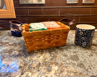 May include: A small woven basket with leather handles filled with tea bags. The basket is sitting on a granite countertop next to a blue and white patterned mug.