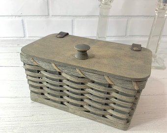 May include: A gray rectangular woven basket with a matching wooden lid. The lid has a small, round knob and is secured with leather straps. The basket is made of woven strips of wood and has a rustic, handmade appearance.