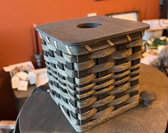 May include: A square, black and gray woven basket with a wooden lid and a small hole in the center. The basket is sitting on a gray wooden surface.