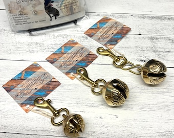 May include: Three gold-colored dog bells with a decorative design. Each bell is attached to a silver swivel clasp. The bells are on a white wooden surface. The text on the card reads: "Amish Made Doggie Hiking Bell".