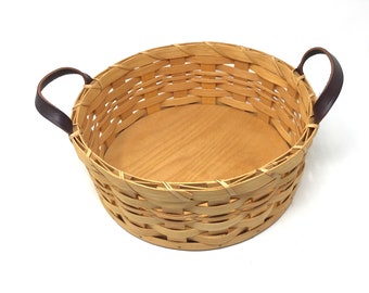 May include: A round woven basket with leather handles. The basket is made of natural wood and has a light brown color. The basket is empty.