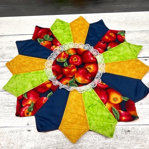 Peut inclure: Un napperon rond avec un motif en étoile. Le napperon est fait de tissu avec un schéma de couleurs vert, jaune et bleu. Le centre du napperon présente un cercle de dentelle blanche avec un imprimé de pommes rouges.