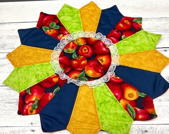 May include: A round table topper with a starburst design. The topper is made of fabric with a green, yellow, and blue color scheme. The center of the topper features a white lace circle with a red apple print.