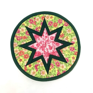 Puede incluir: Un posavasos redondo de tela verde con estampado floral rosa y verde. El centro del posavasos presenta una gran estrella de ocho puntas con estampado floral rosa.