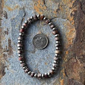 May include: A beaded bracelet with a mix of metallic and speckled beads. The bracelet is displayed on a textured, multi-colored stone surface. A U.S. dime is next to the bracelet for size reference.