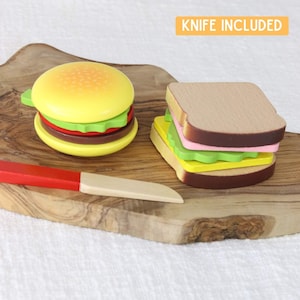Puede incluir: Un juego de madera con una hamburguesa y un sándwich en una tabla de cortar de madera. Se incluye un cuchillo de plástico rojo y blanco. "KNIFE INCLUDED" está escrito en una etiqueta amarilla.