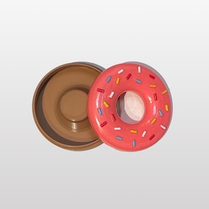 Könnte beinhalten: Ein rosafarbener Behälter in Donutform mit bunten Streuseln, der geöffnet ist und einen braunen Deckel zeigt.