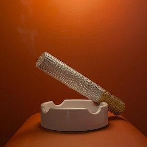 May include: A decorative cigarette with a silver textured body and a gold tip rests in a white ashtray. Wisps of smoke rise against an orange background, suggesting a smoking theme.