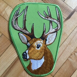 Può includere: Toppa ricamata raffigurante un cervo con grandi corna. Il cervo ha un mantello marrone, un muso bianco, su uno sfondo verde chiaro. La toppa ha un bordo verde, ideale per abbigliamento o accessori.