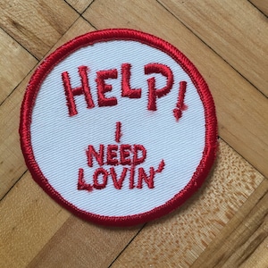 May include: White fabric patch with red embroidered text that reads "HELP! I NEED LOVIN'" with a red stitched border.