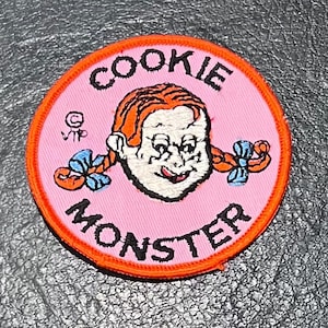 May include: A pink and orange embroidered patch with the text "Cookie Monster" in black lettering. A cartoon image of Cookie Monster with red hair and blue bows is in the center of the patch.