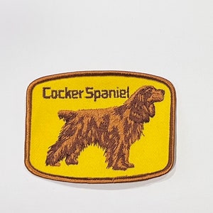 Puede incluir: Parche bordado con un perro Cocker Spaniel de perfil. El perro es marrón con un aspecto texturizado, sobre un fondo amarillo. Las palabras "Cocker Spaniel" están bordadas encima del perro en color marrón oscuro. El parche tiene un borde marrón.