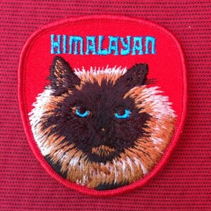 Può includere: Una toppa ricamata rossa con il viso di un gatto himalayano e la parola "Himalayan" in lettere blu.