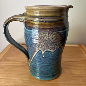 May include: A ceramic pitcher featuring a tall, cylindrical body with a curved handle and a small pouring spout. The exterior displays a reactive glaze with a blend of deep blue, earthy brown, and tan tones, characterized by a textured finish.