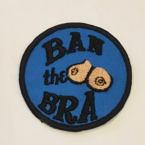 May include: Circular patch with a blue background and a black border. The patch displays the words "BAN the BRA" in black, decorative lettering. Two tan-coloured, embroidered breast shapes are also present.