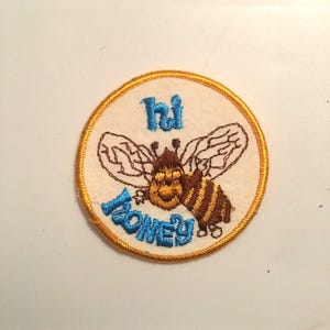 Könnte beinhalten: Ein runder, weißer gestickter Aufnäher mit gelbem Rand. Der Aufnäher zeigt eine Cartoon-Biene mit braunen und gelben Streifen und dem Text "hi honey" in Blau.