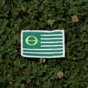 Könnte beinhalten: Ein gestickter Aufnäher in Grün und Weiß mit einem gelben Kreis, in dem sich ein grüner Buchstabe "O" befindet. Der Aufnäher ist eine stilisierte amerikanische Flagge mit grünem Hintergrund und weißen Streifen.