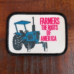 May include: Rectangular patch with a black border featuring a blue tractor illustration. The text "FARMERS THE ROOTS OF AMERICA" is printed in pink. The patch is on a wooden surface.