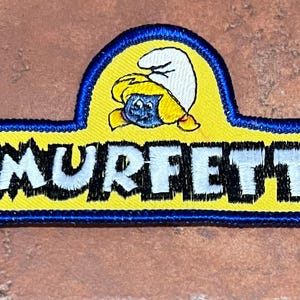 May include: Embroidered patch with the word "SMURFETTE" in white on a yellow background. A blue border surrounds the yellow rectangle and the top section, which features a cartoon Smurfette character.