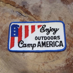 Peut inclure: Patch brodé blanc avec un motif de drapeau américain rouge, blanc et bleu. Le texte "Enjoy Outdoors Camp AMERICA" est brodé en noir.