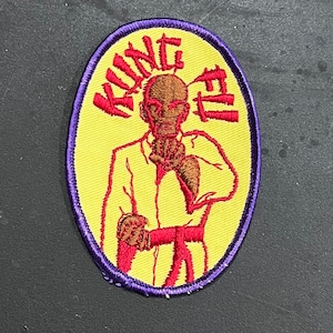 Peut inclure: Un écusson brodé de forme ovale jaune avec une bordure violette. L'écusson représente un personnage de dessin animé en uniforme de karaté rouge avec une ceinture rouge, effectuant une pose de karaté. Le texte "KING FU" est brodé en rouge autour de la tête du personnage.