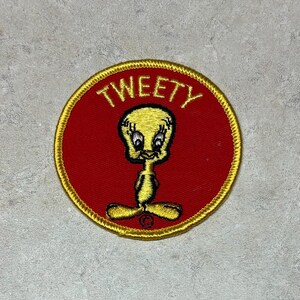 May include: A circular patch featuring Tweety Bird from Looney Tunes. The patch has a red background with a gold border. The word "TWEETY" is embroidered in gold above the yellow cartoon bird.