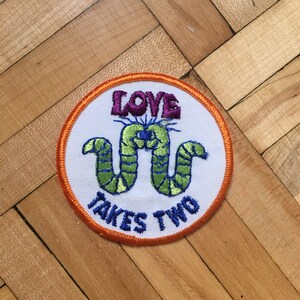 Peut inclure: Un écusson brodé blanc avec une bordure orange. L'écusson représente deux vers verts entrelacés avec le texte "LOVE TAKES TWO" en lettres violettes et bleues.