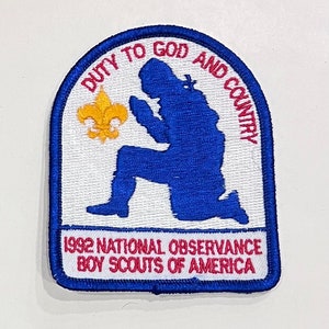 Peut inclure: Un écusson rectangulaire avec une bordure bleue et un fond blanc. L'écusson présente une silhouette bleue d'une personne agenouillée en prière, un fleur-de-lis doré et le texte "DUTY TO GOD AND COUNTRY". Le texte du bas indique "1992 NATIONAL OBSERVANCE BOY SCOUTS OF AMERICA".