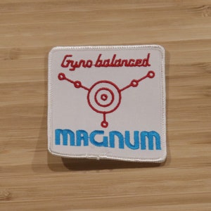 Può includere: Toppa ricamata bianca con cuciture rosse e blu. La toppa presenta un bersaglio stilizzato con tre linee che si estendono da esso. Il testo "Gyro balanced" è sopra il bersaglio e "MAGNUM" è sotto il bersaglio.