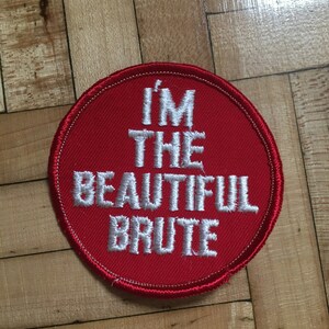 May include: Red round embroidered patch with white stitching. The text on the patch reads "I'm the beautiful brute."