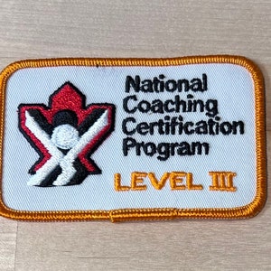 May include: White embroidered patch with a red, black, and white logo of a stylized maple leaf with a person inside. The text "National Coaching Certification Program" and "LEVEL III" are embroidered in black and orange.