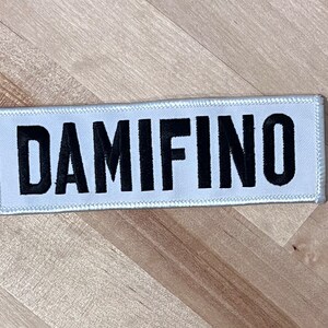 May include: White fabric patch with black embroidered text that reads "DAMIFINO".