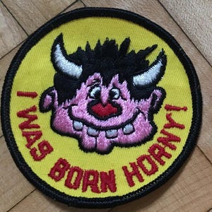 May include: A yellow and black round patch with a pink cartoon monster with horns and the text "I was born horney!" in red.