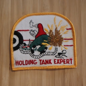 Könnte beinhalten: Ein weißer gestickter Aufnäher mit gelbem Rand. Der Aufnäher zeigt eine Comic-Illustration einer Person, die einen explodierenden Panzer hält. Der Text "HOLDING TANK EXPERT" ist in Schwarz unter der Illustration geschrieben.