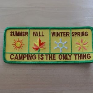 May include: A yellow embroidered patch with green trim. The patch has four sections, each representing a season: summer, fall, winter, and spring. Each section has an embroidered image representing the season. The text "Camping is the only thing" is embroidered in brown on the bottom of the patch.